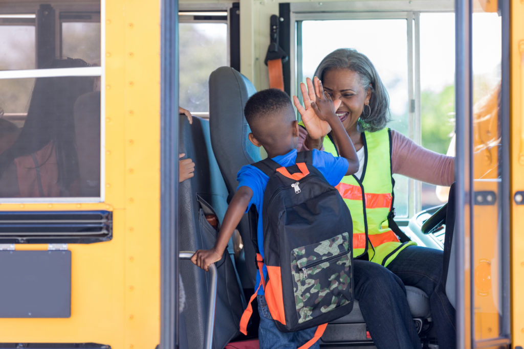A female school bus driver high-fives a new student to make him feel welcome as he enters the bus.